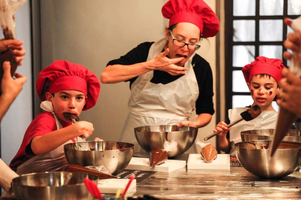 Actividades de verano Interlaken Parte 3 5 Niños y adultos en el taller de chocolate, juntos con gorros de cocinero rojos y comiendo chocolate en la cocina.