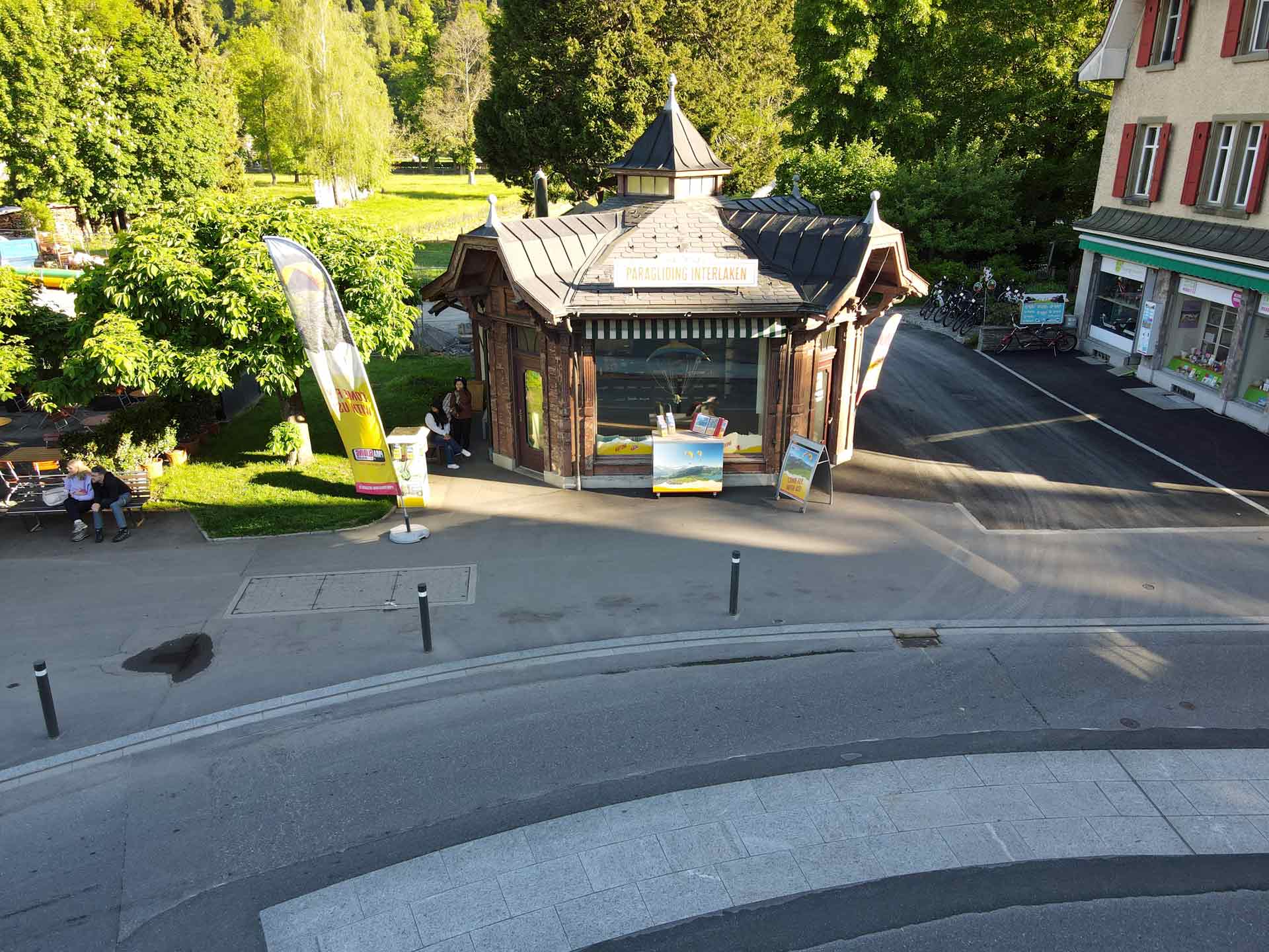 Punto de encuentro en Interlaken 1 Historischer Kiosk in Interlaken umgeben von Grün und Straßen, mit Werbeflagge bei Sonnenschein.