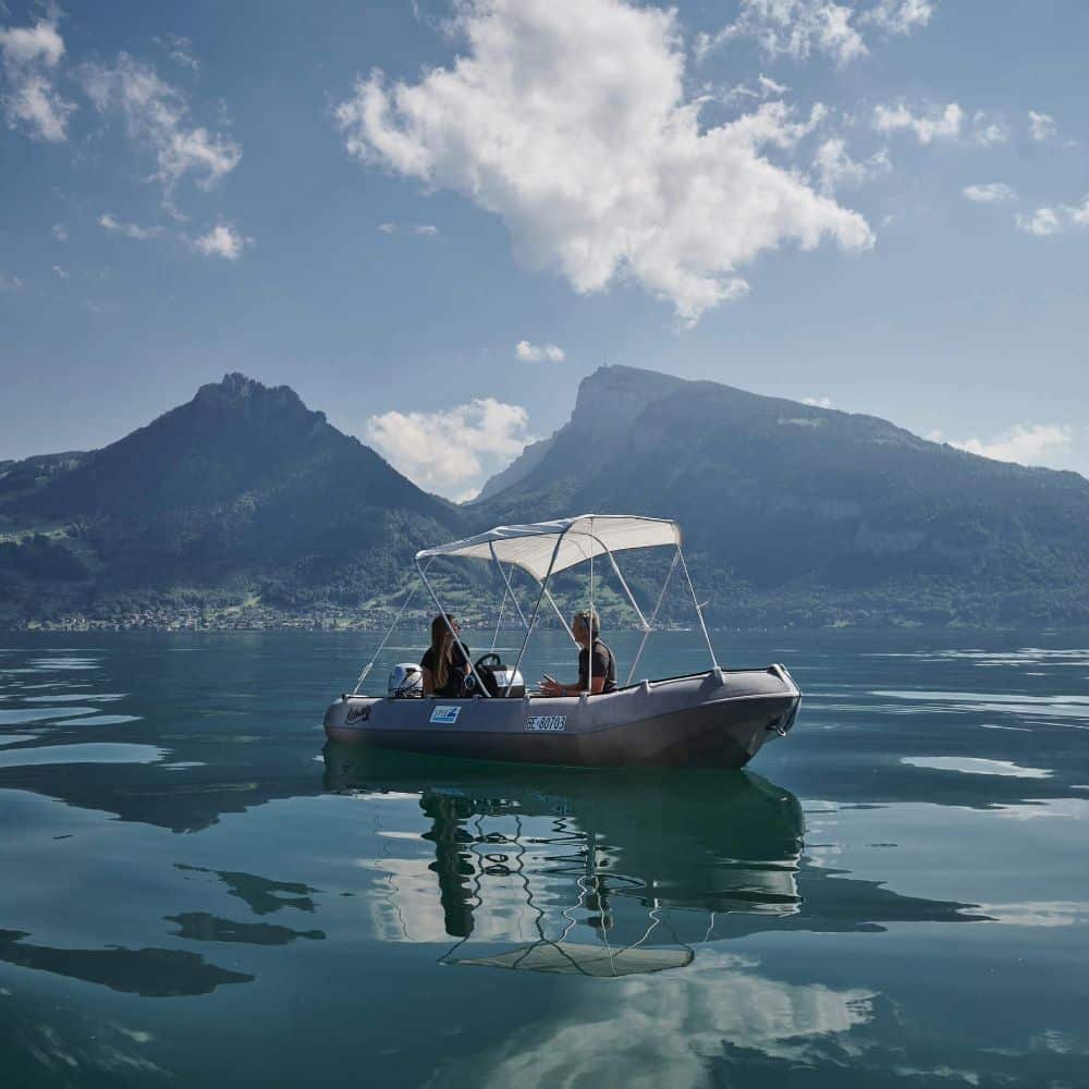 Bateau sur un lac entouré de montagnes sous un ciel bleu, avec deux personnes à bord contemplant le paysage.