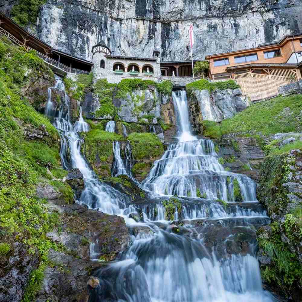 Chalet pittoresque sur une falaise avec cascade, entouré de verdure alpine, drapeau suisse flottant.