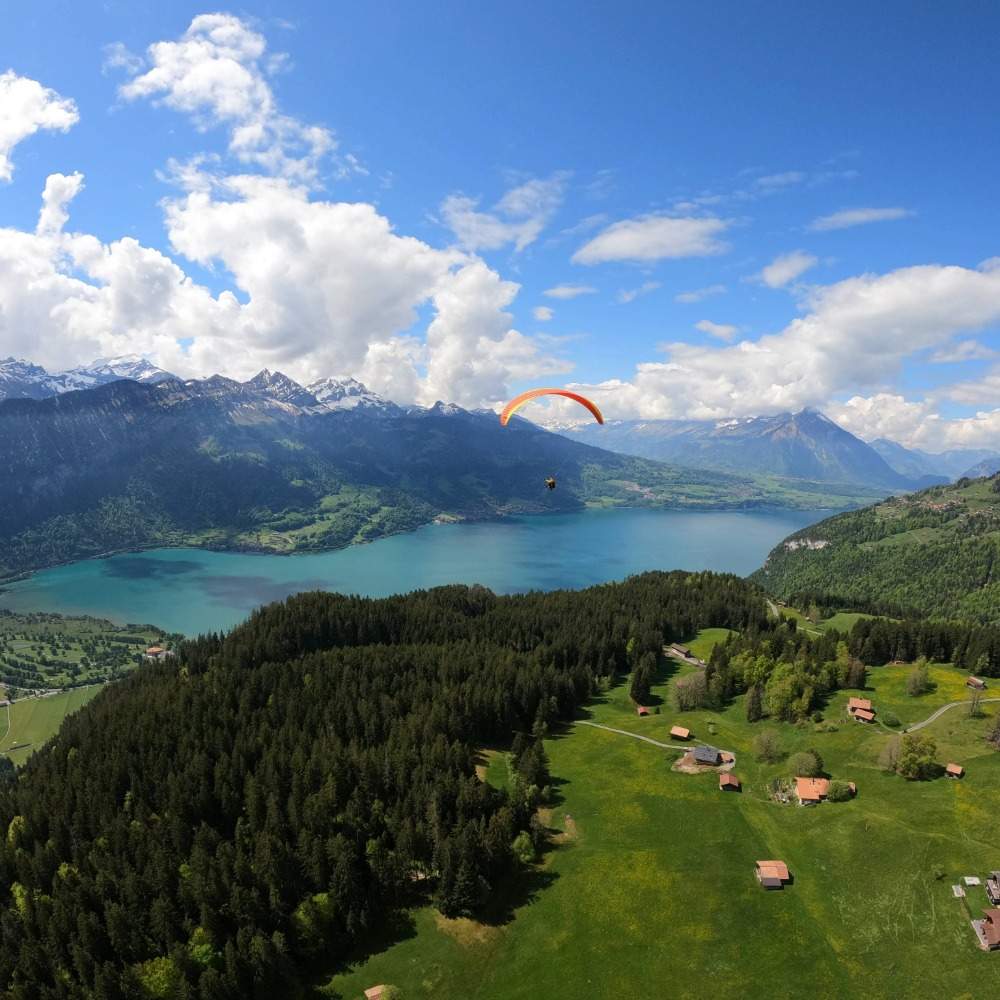 Un parapente survole un lac alpin entouré de montagnes verdoyantes et de forêts, sous un ciel bleu parsemé de nuages.