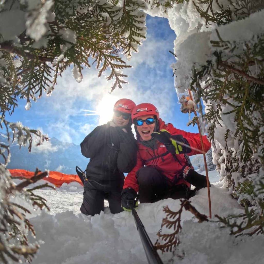 Deux personnes souriantes avec des casques rouges et des équipements de ski, posant dans la neige sous un ciel ensoleillé.