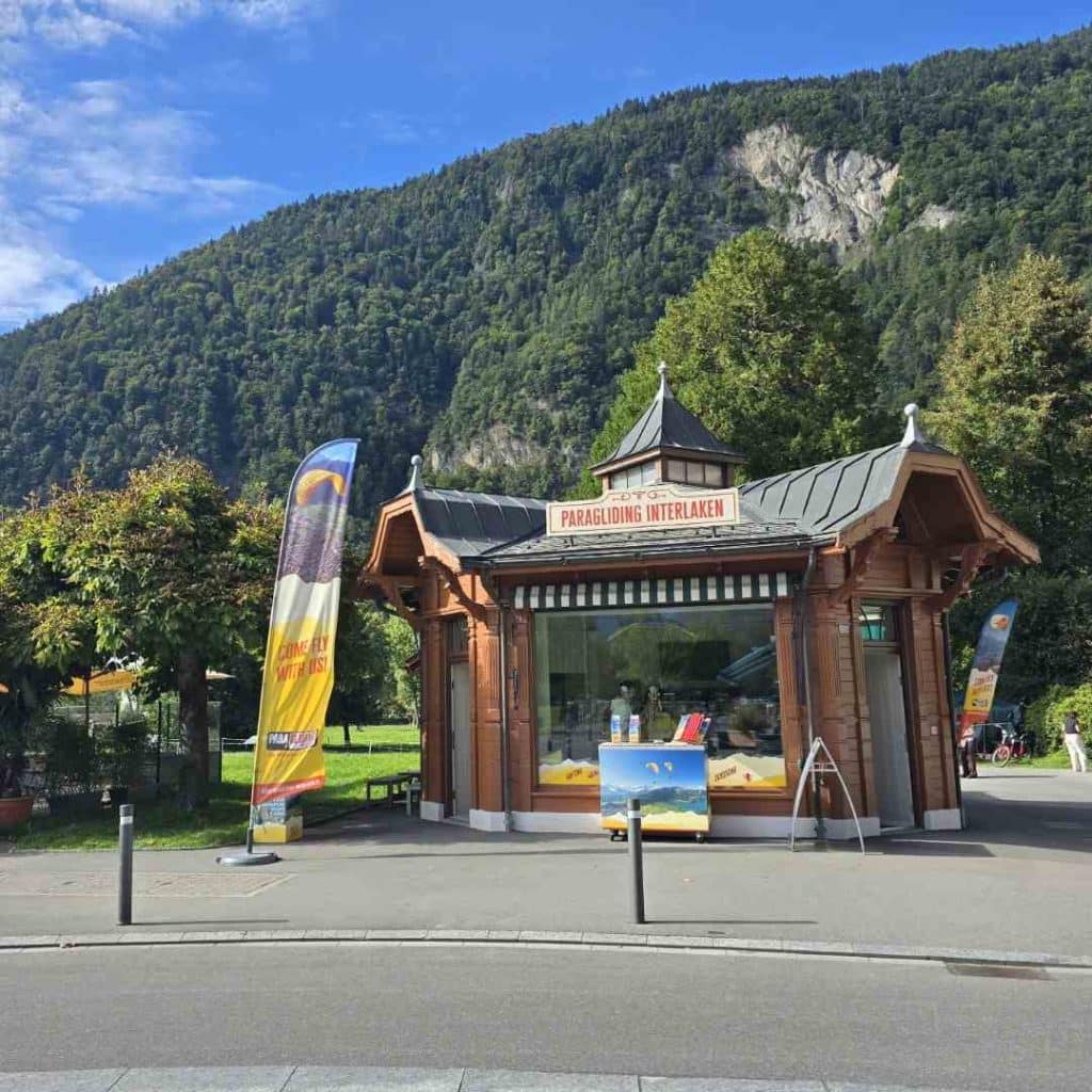 Boutique de parapente en bois à Interlaken, entourée de montagnes verdoyantes et ciel bleu en arrière-plan.