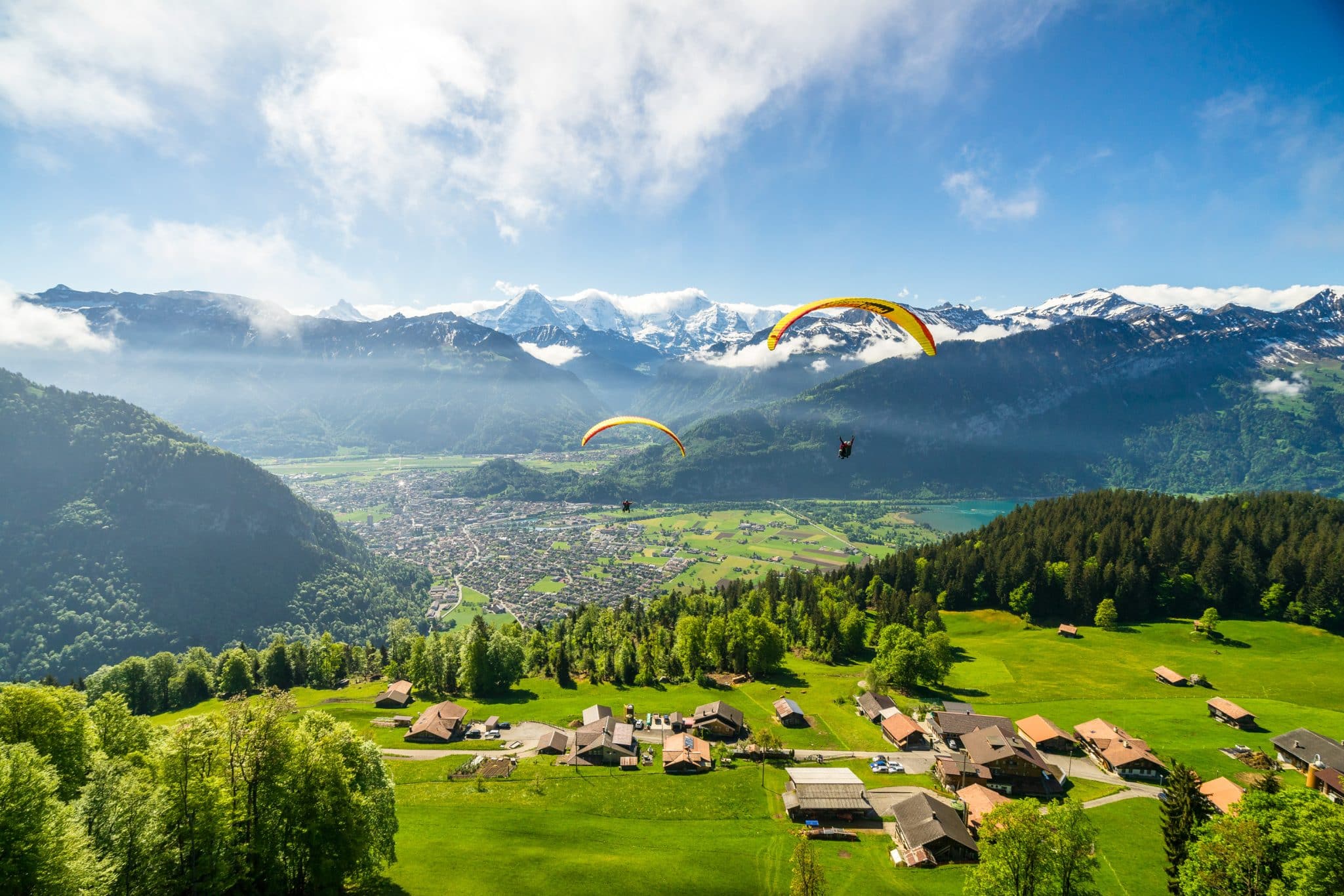 Parapentes survolant un village alpin avec les montagnes enneigées en arrière-plan par une journée ensoleillée.