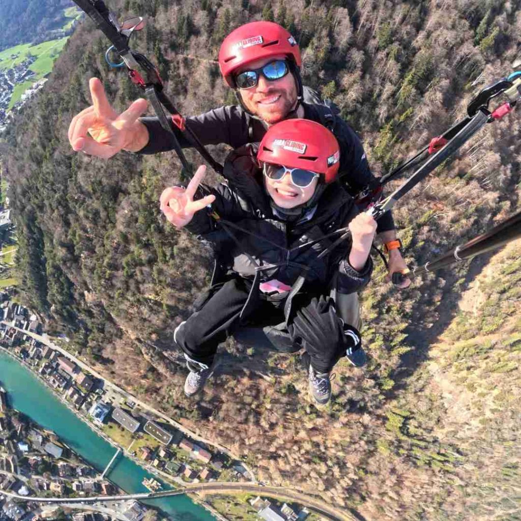 Deux personnes en parapente survolant un paysage montagneux et une ville, souriantes avec des casques rouges.