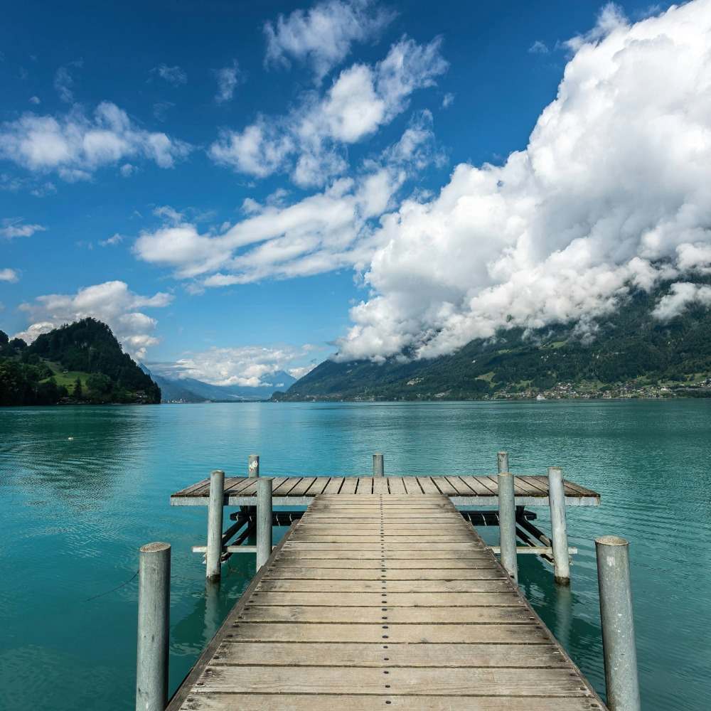 Quai en bois sur un lac alpin turquoise sous un ciel bleu avec des montagnes et des nuages blancs en arrière-plan.