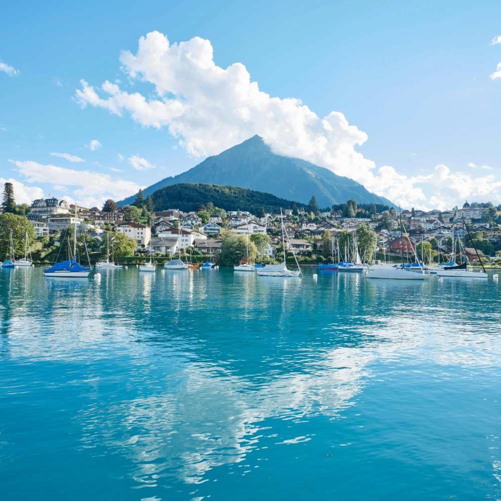 Paysage alpin avec voiliers sur un lac, montagnes et ville pittoresque sous un ciel bleu.