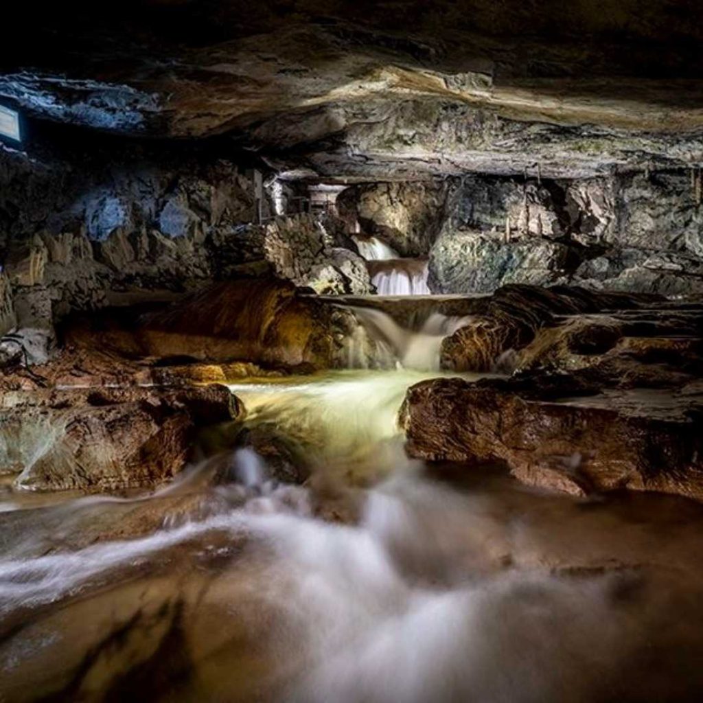 Cascade souterraine illuminée dans une caverne rocheuse, eau fluide courant sur des pierres sombres et brillantes.