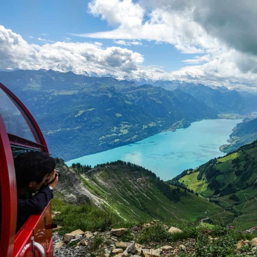 Vue depuis un train rouge sur un lac alpin bleu entouré de montagnes verdoyantes et ciel partiellement nuageux.